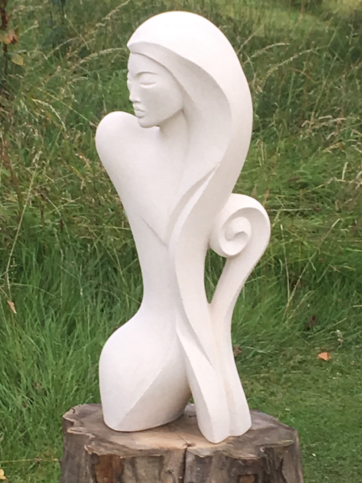 Pale abstract figurative sculpture displayed on a tree stump in a garden